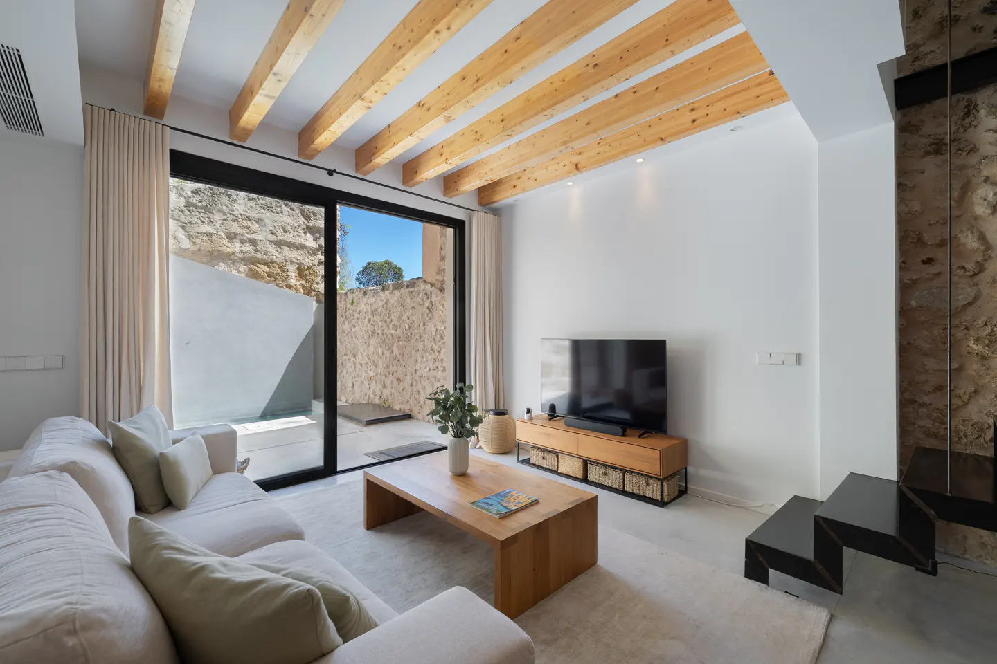 Living room with white sofa, wood coffee table, TV, and sliding glass doors to a patio with stone walls. Wood beams on ceiling.