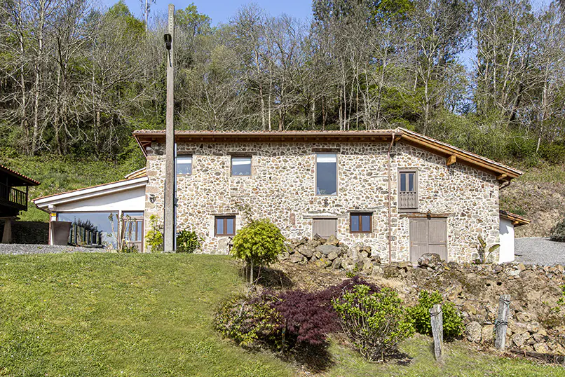 Exterior view of a stone house with a brown roof, set on a green hillside with trees in the background.