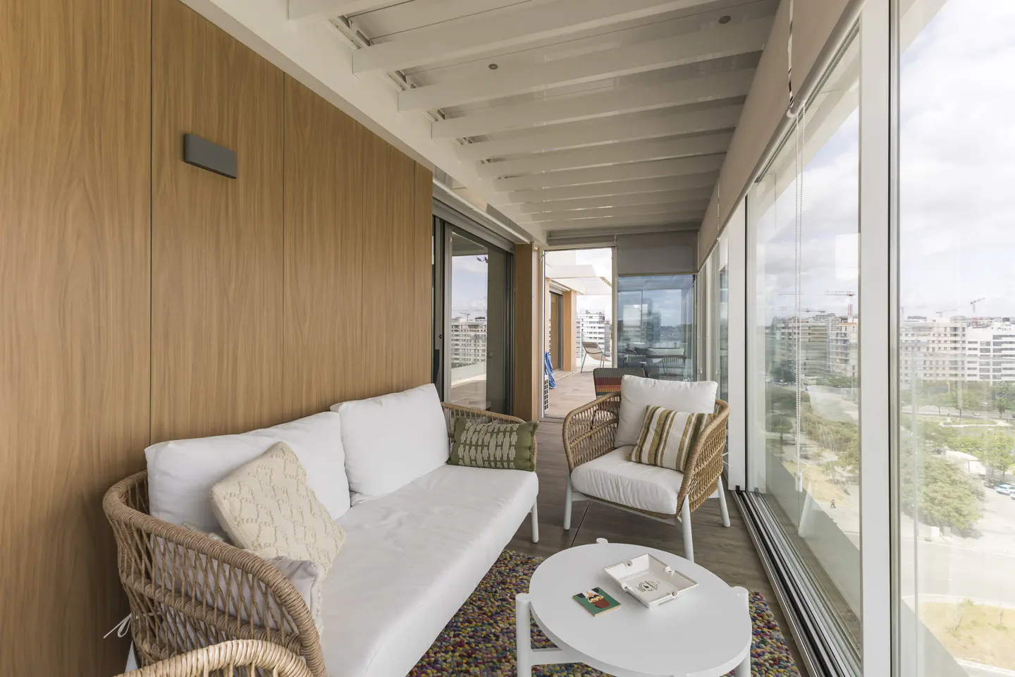 A bright sunroom with a white sofa, wicker chairs, and a white table. Large windows offer a city view.