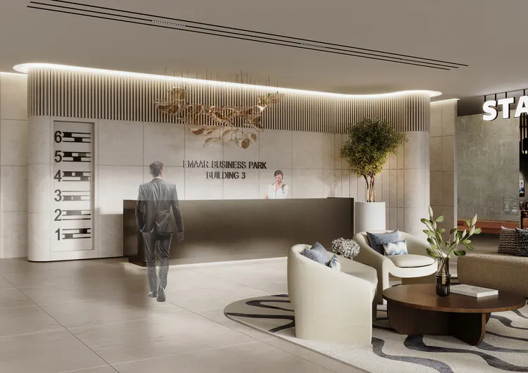 Modern office lobby with a man walking towards the reception desk. Seating area with white chairs and a round coffee table. "Emaar Business Park Building 3" is on the wall.