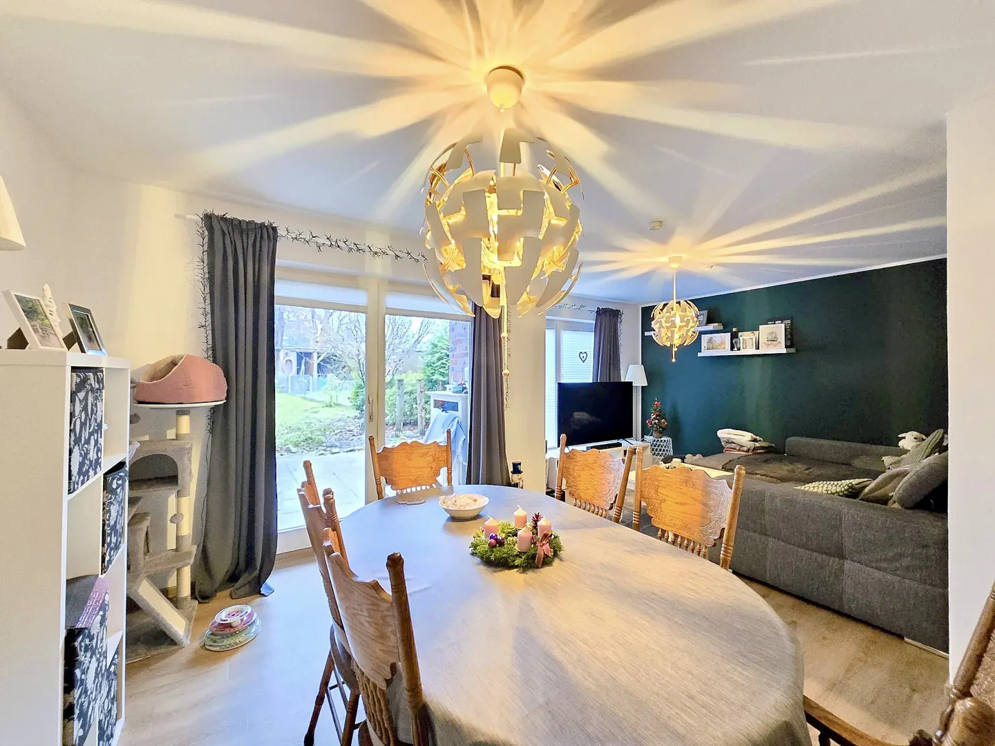 Bright dining room with an oval table, wooden chairs, and a modern chandelier. A gray sofa and TV are visible in the background.