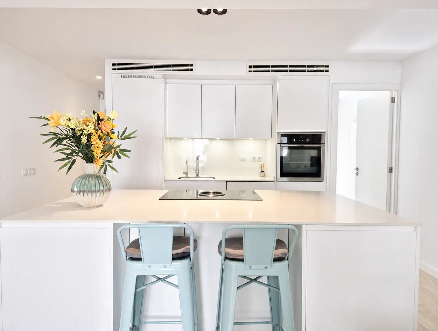 Bright, modern kitchen with white cabinets, countertops, and appliances. Two blue bar stools face the island. A vase of yellow flowers adds a pop of color.