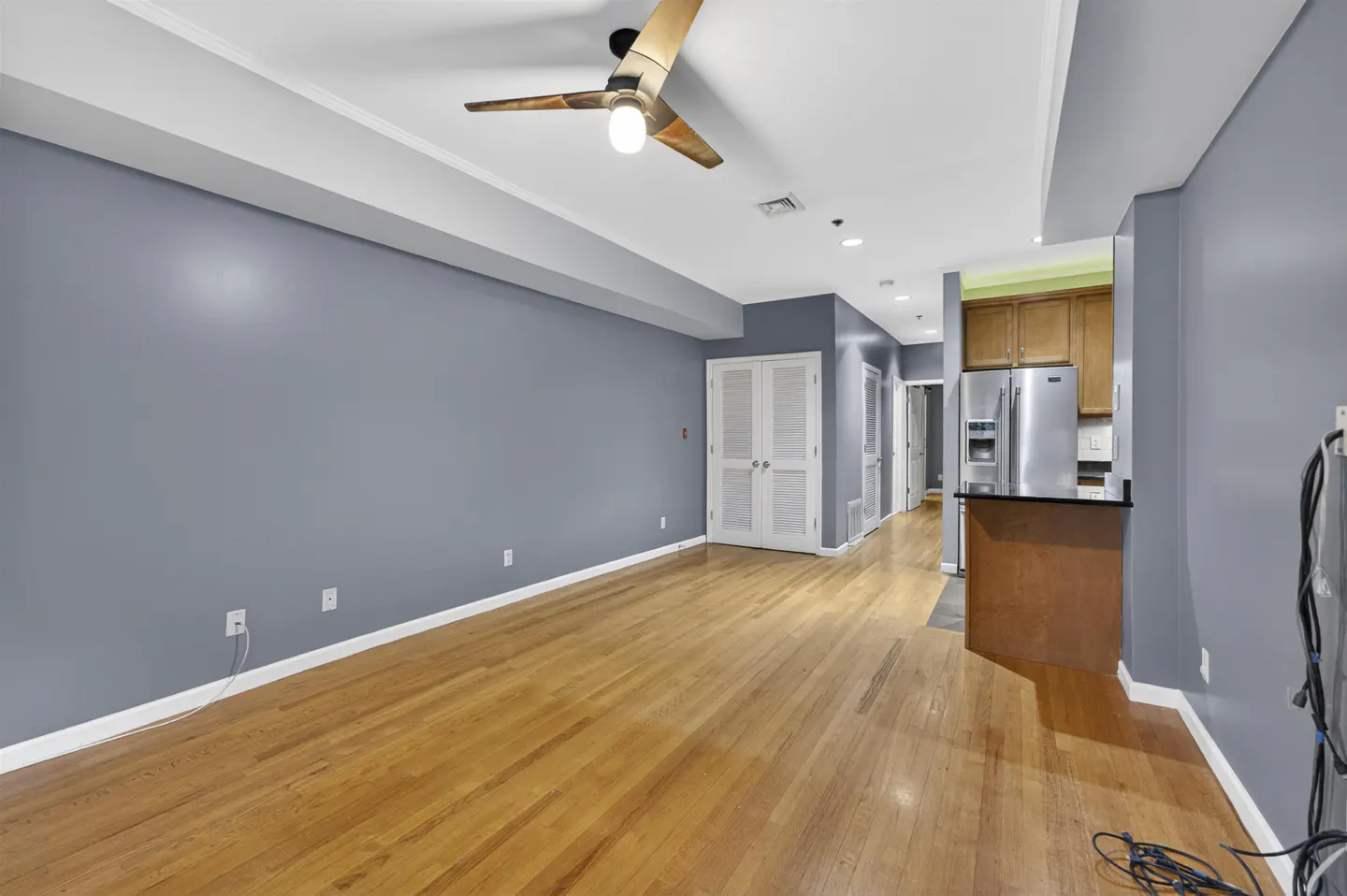 Spacious living area with hardwood floors, blue-grey walls, a ceiling fan, and an open kitchen with stainless appliances.