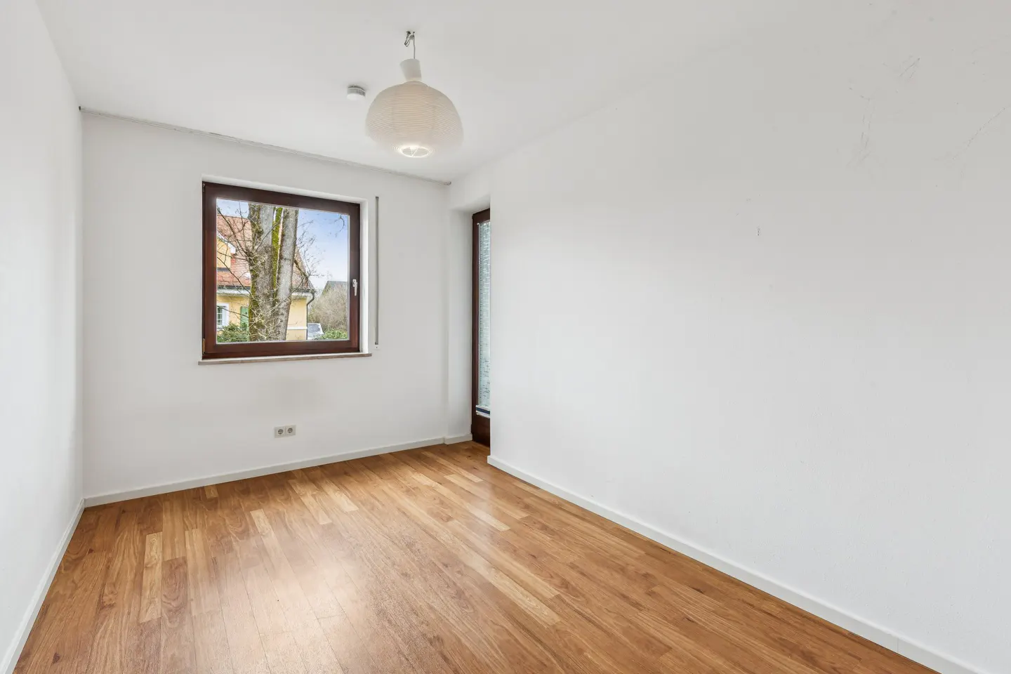 Bright, empty room with white walls, wood floor, and a window showing a tree and houses. A modern light fixture hangs from the ceiling.