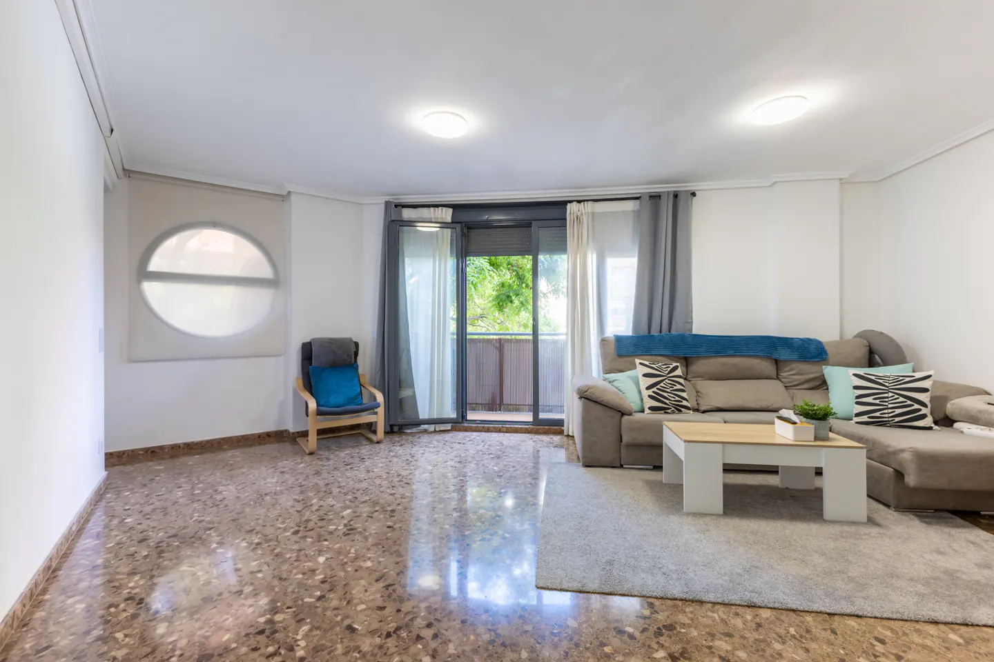 Bright living room with a gray sofa, white coffee table, and a chair near a round window and balcony door.