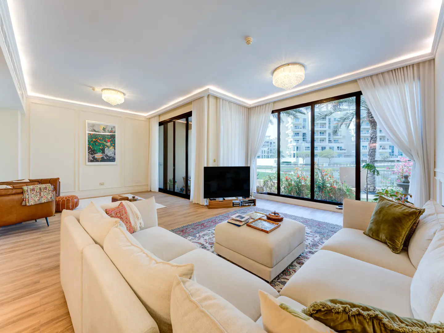 Bright living room with a white sectional sofa, wood floors, and large windows with sheer curtains overlooking a garden.