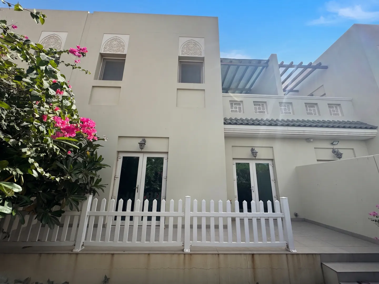 Two-story beige house with white trim, windows, and doors. A white picket fence lines the patio. Pink flowers are on the left.