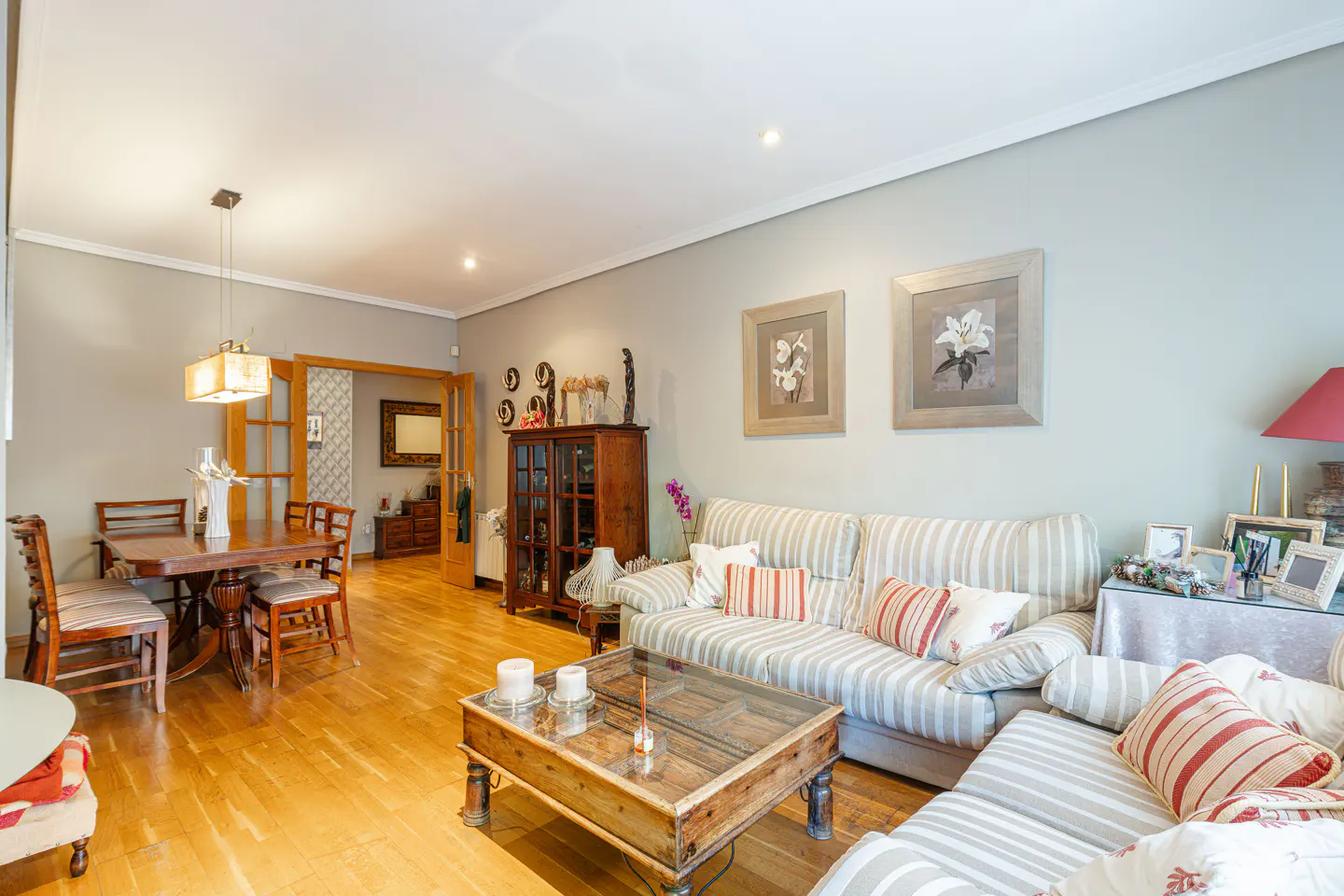 Living room with wood floors, striped sofa, glass coffee table, dining table, and framed floral art.