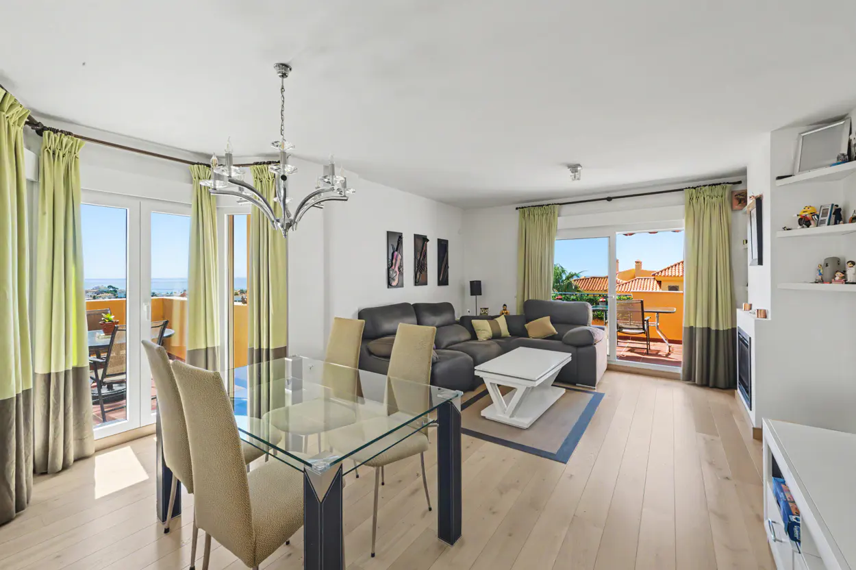 Bright living room with a glass dining table, gray sofa, and balcony access with ocean views.