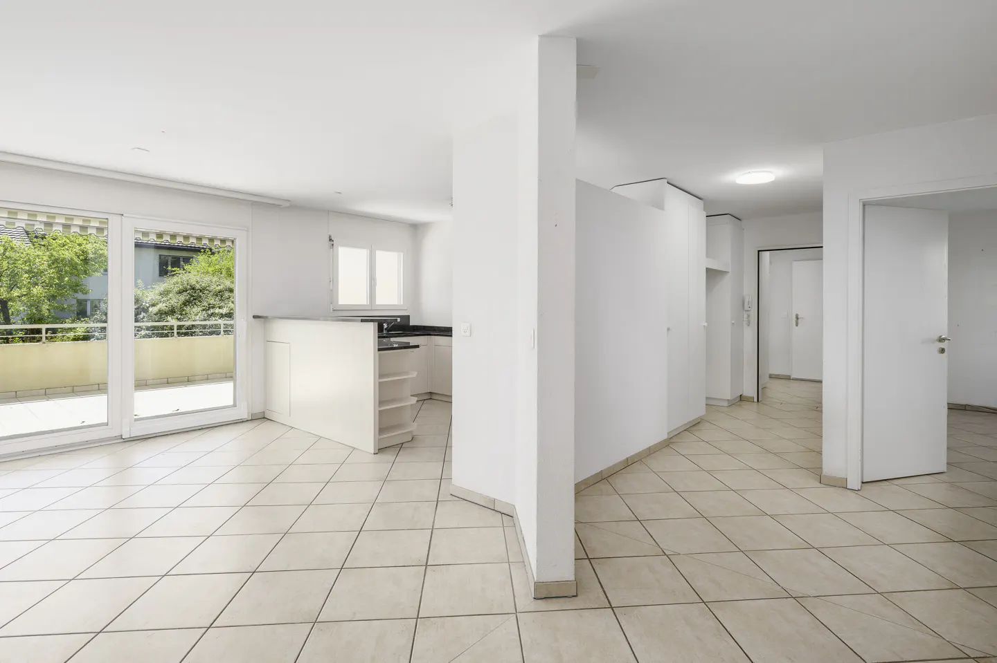 Bright, open-plan apartment with white walls, tiled floors, a small kitchen, and glass doors leading to a balcony.