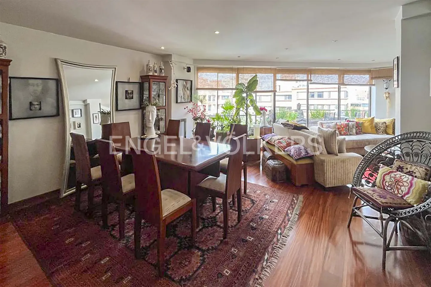 A bright living and dining area with a wooden table, chairs, and a patterned rug. A sofa and wicker chair are near a window with a city view.