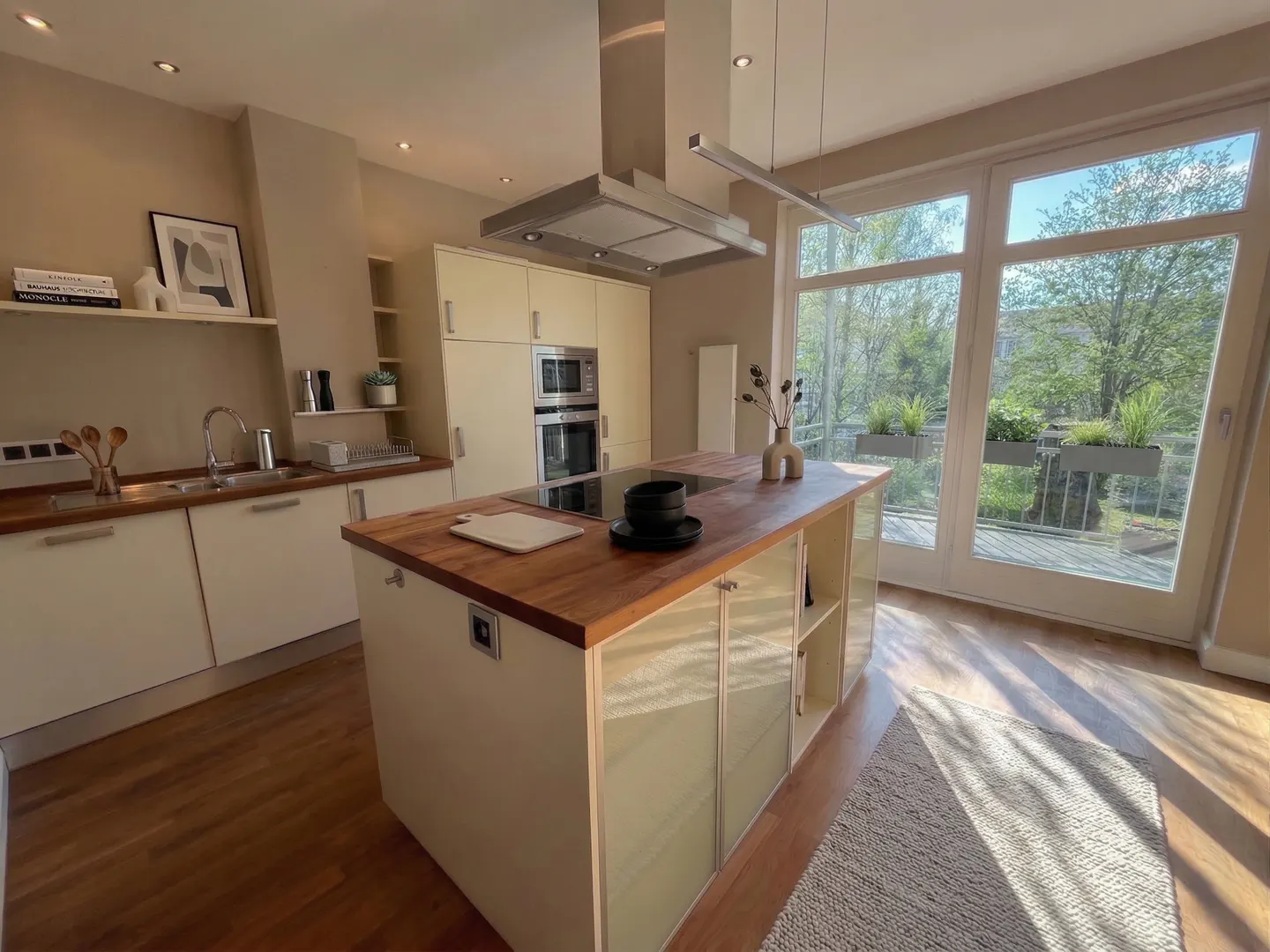 Bright kitchen with wood floors, cream cabinets, and a wood-topped island. Large windows overlook greenery.