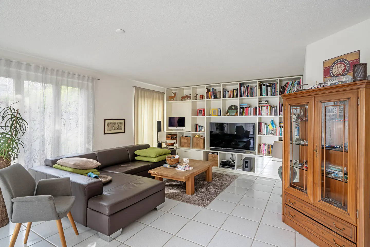 Living room with brown leather sectional, gray chair, wood coffee table, and white built-in shelving unit with TV and books.
