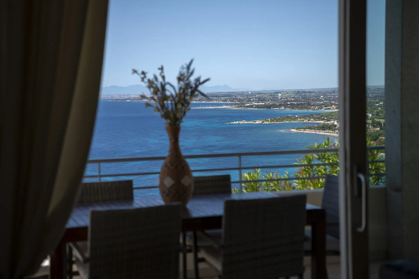 Ocean view from a dining room. A vase of flowers sits on a dark wood table with chairs. The blue ocean is visible through a sliding glass door.