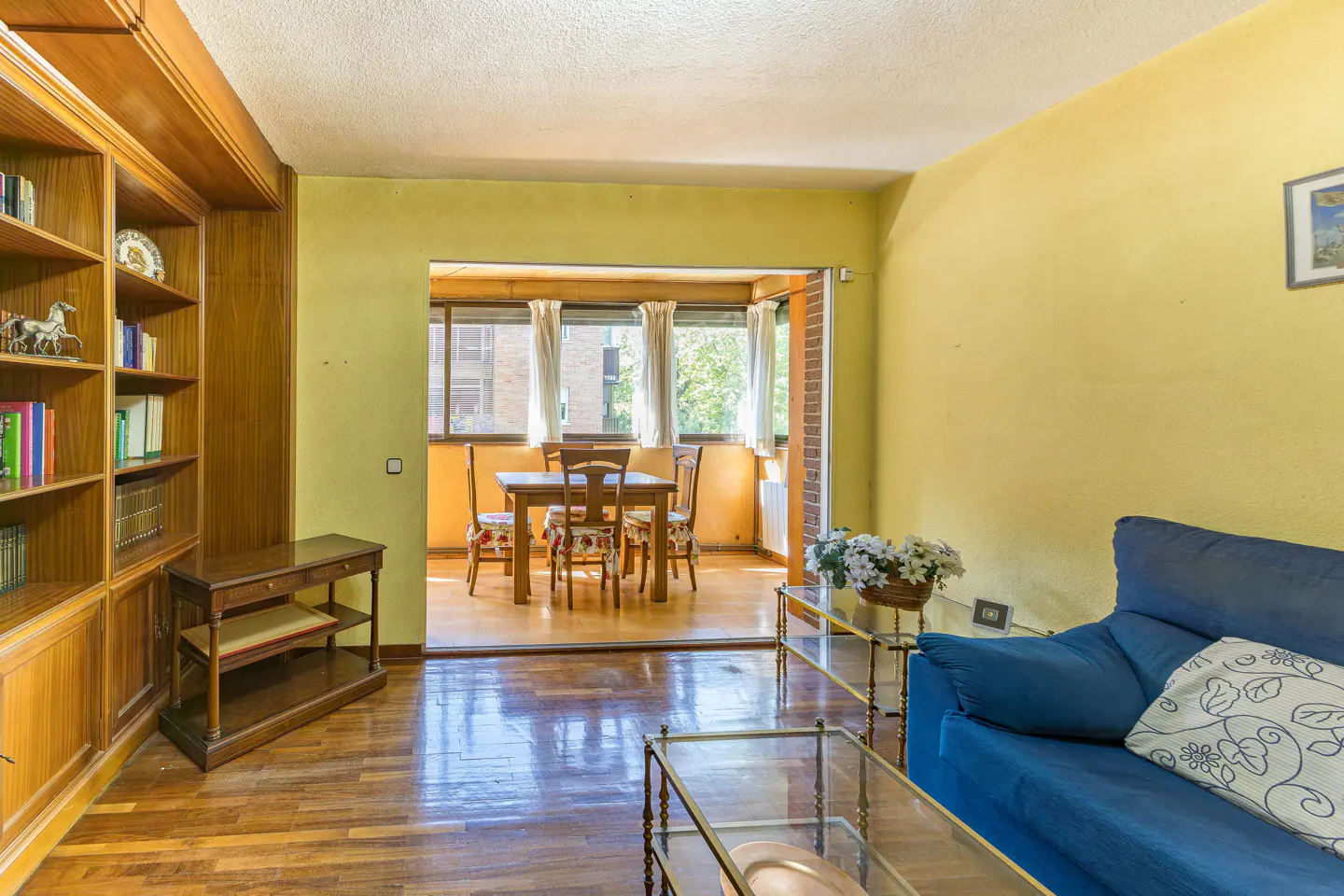 A living room with yellow walls, wood floors, and a blue sofa. A dining table is visible through an open doorway. A wooden bookcase is on the left.