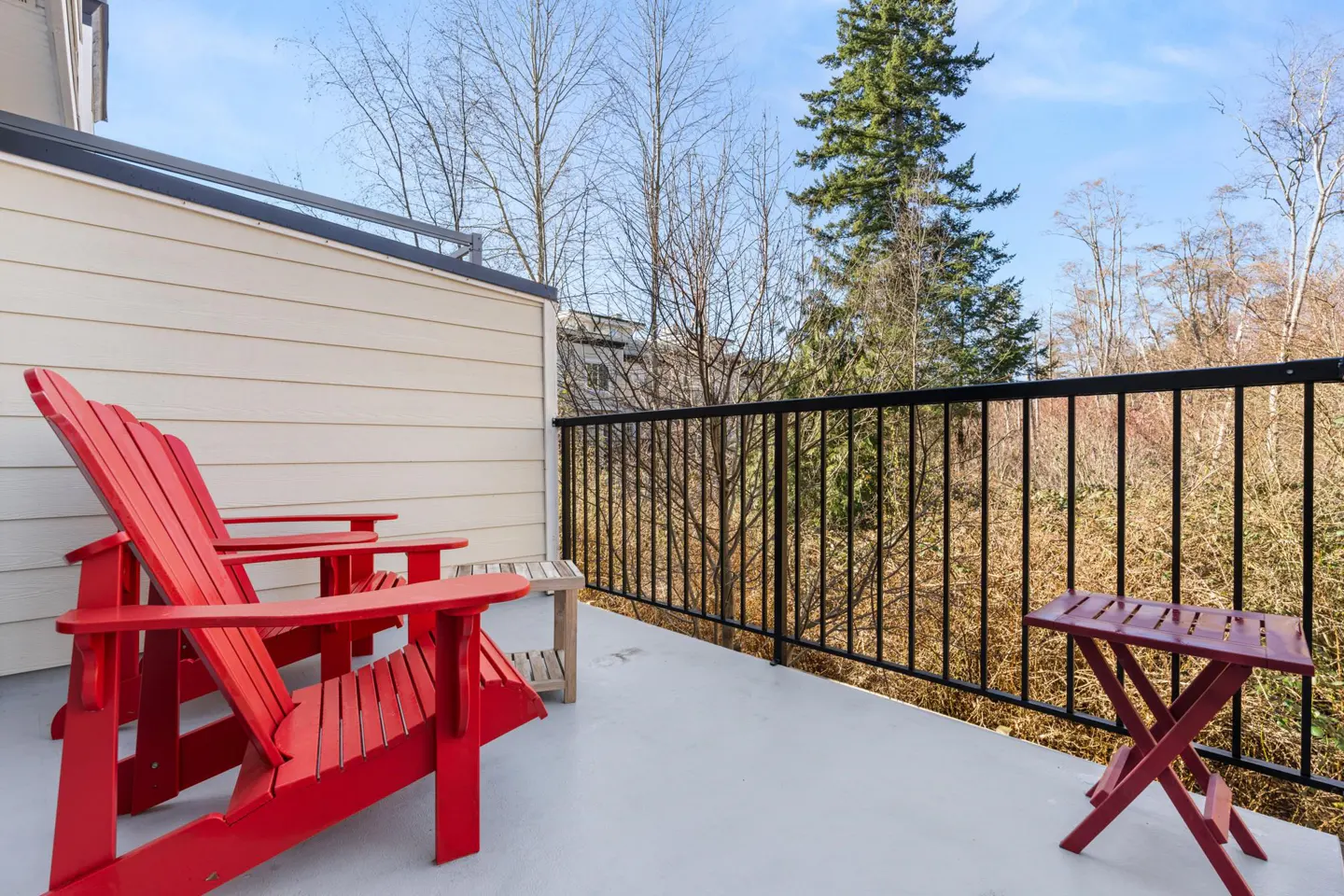 Outdoor patio with two red Adirondack chairs, a small table, and a black metal railing overlooking trees.
