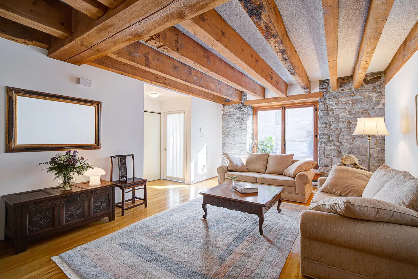 Living room with exposed wood beams, stone wall, and hardwood floors. Two beige sofas face a dark wood coffee table. A large rug covers the floor.