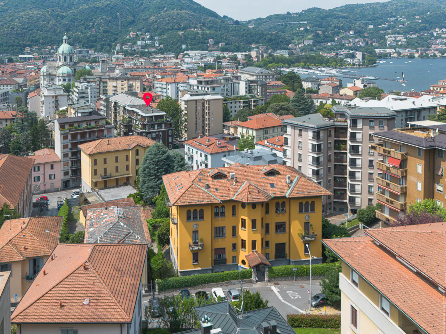 Aerial view of a yellow building with a red tile roof in a dense Italian city, with a red location pin marking a property.