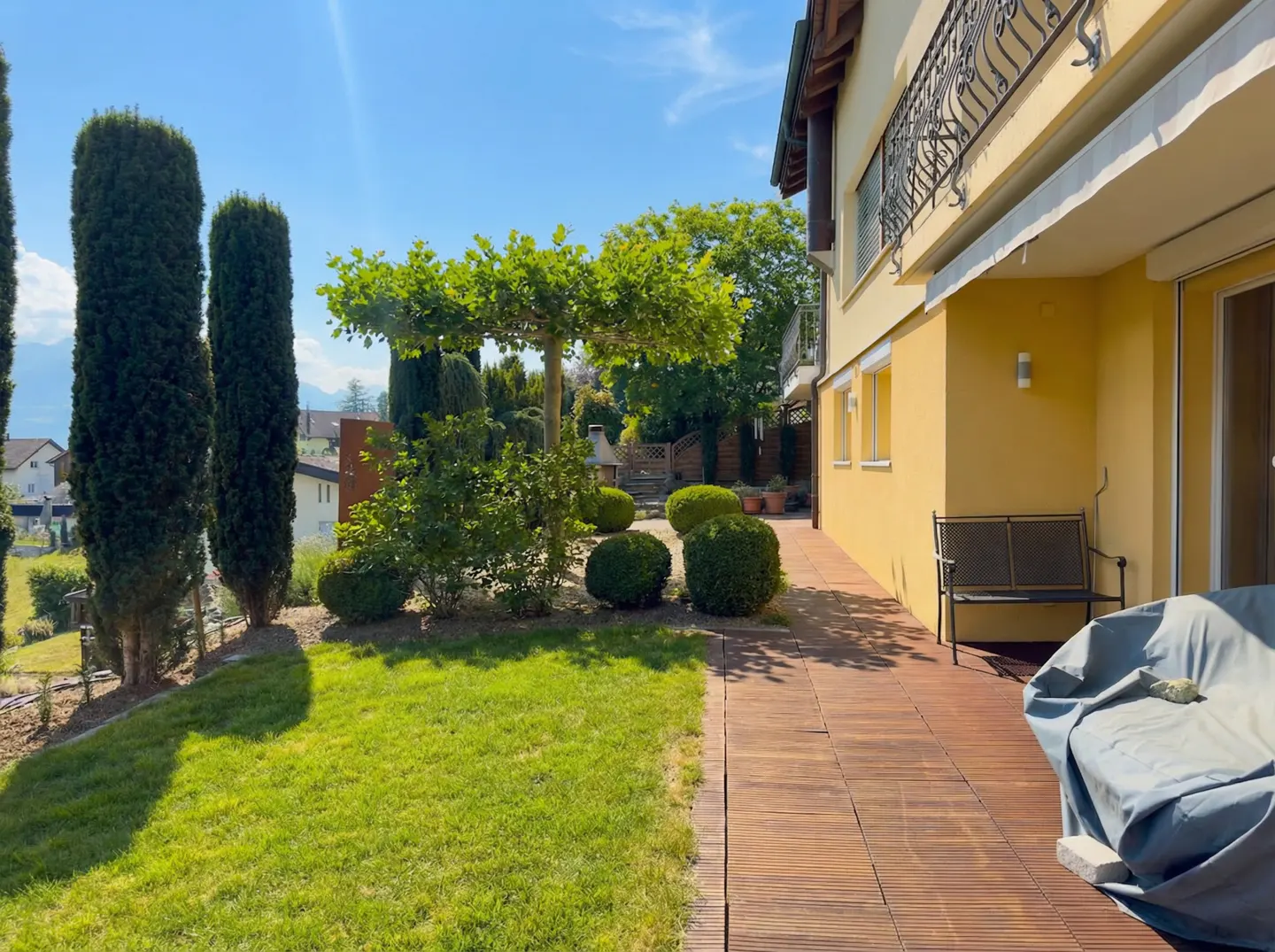 Sunny backyard with green lawn, trimmed bushes, and tall trees. A yellow house with a balcony overlooks a wooden patio with a bench.