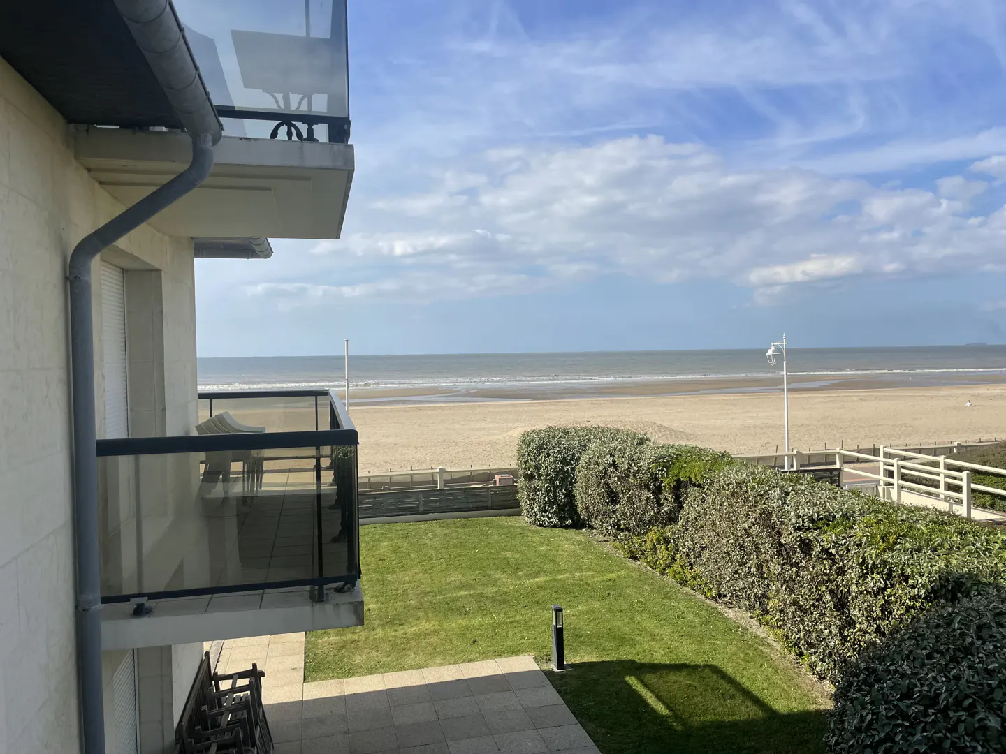 View from a balcony overlooking a green lawn, bushes, sandy beach, and ocean under a cloudy blue sky.