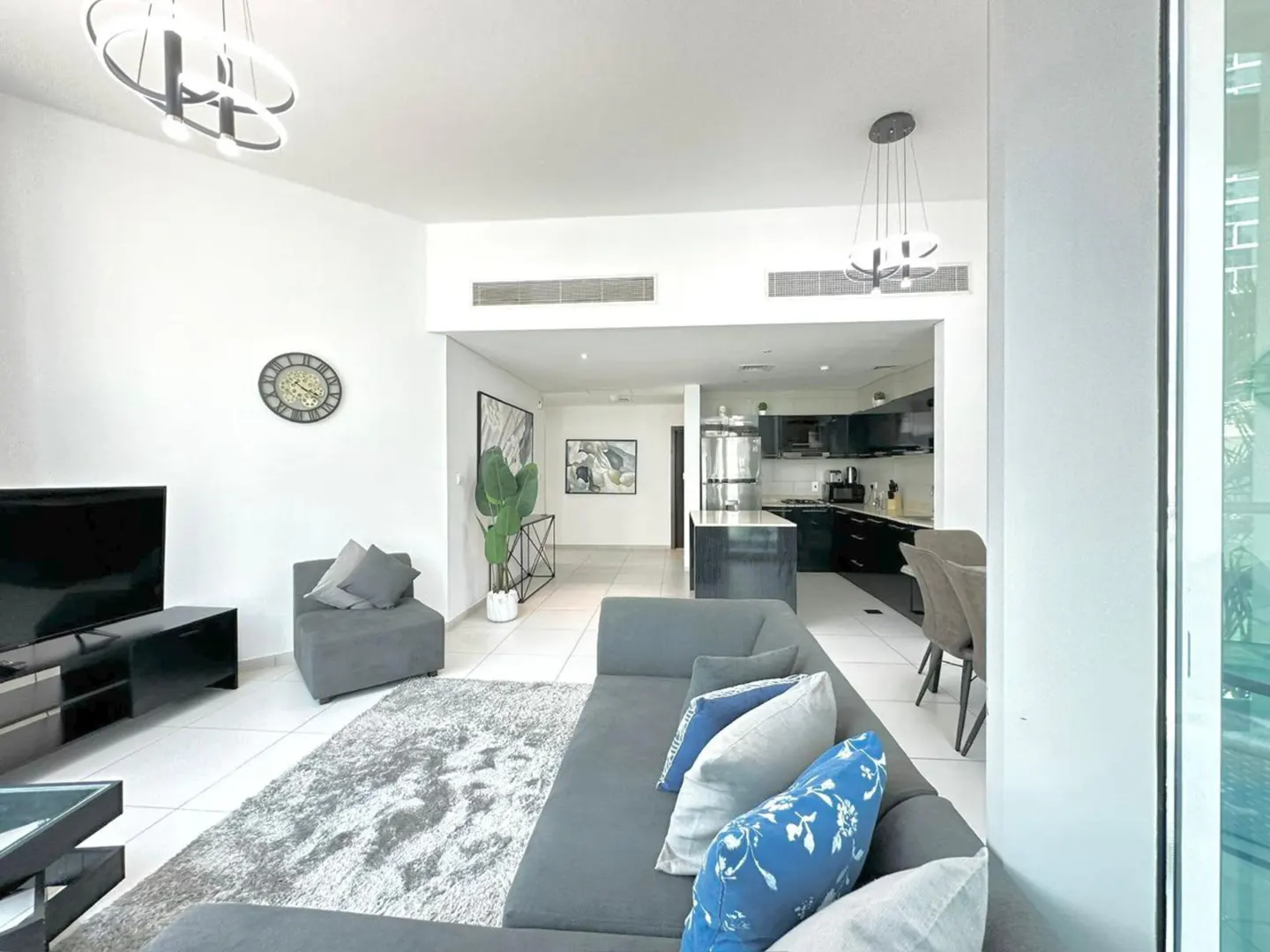 A modern living room with a gray sofa, blue pillows, a TV, and an open kitchen with black cabinets.