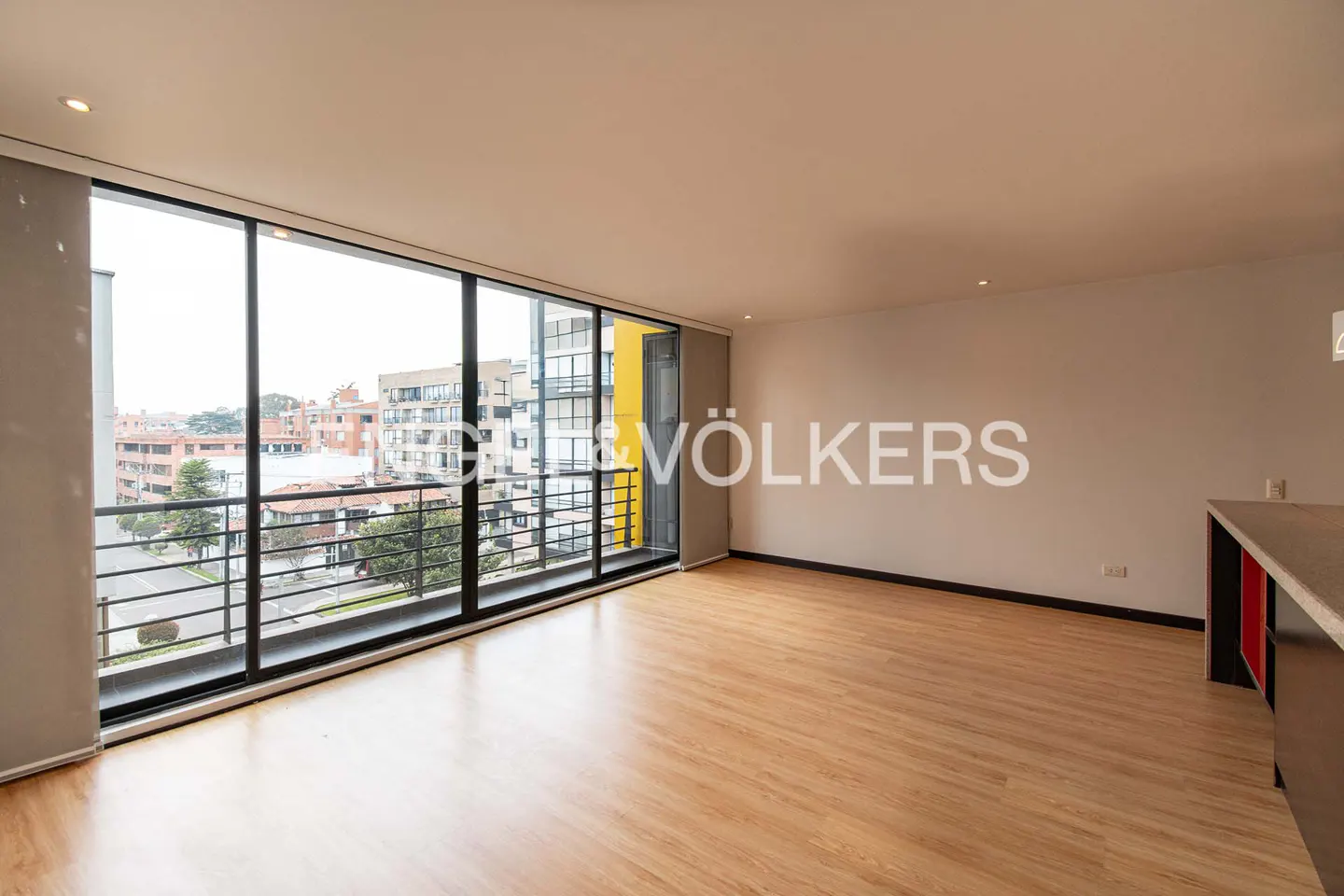 Bright, empty room with wood floors and a large window showing a city view. A kitchen counter is visible on the right.