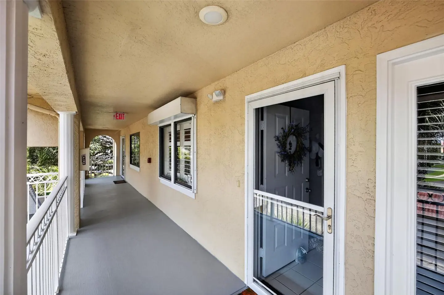 A covered outdoor walkway with beige stucco walls, a gray floor, white railings, and a white screen door with a wreath.