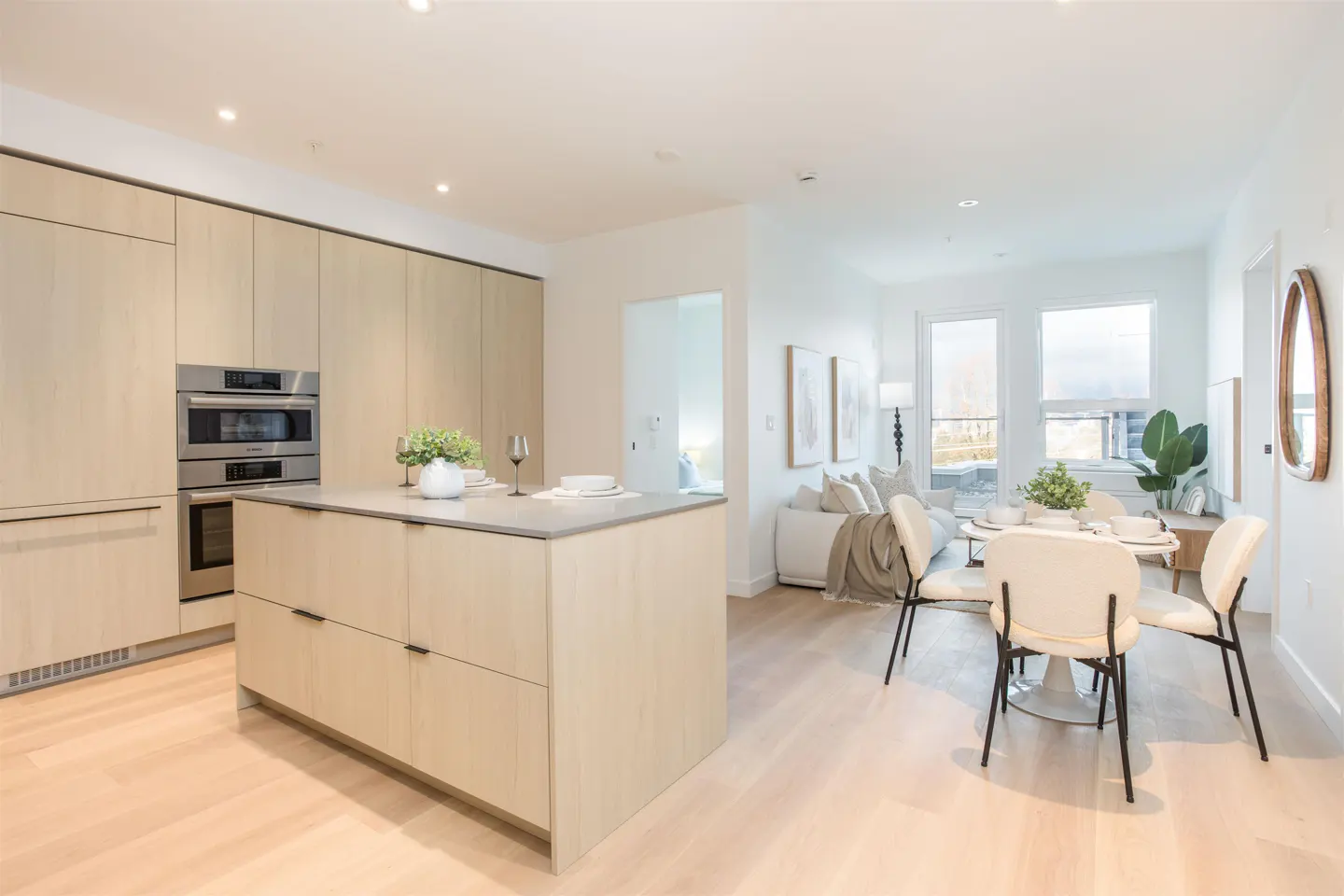 Bright, open-concept kitchen and living area with light wood cabinets, island, dining table with white chairs, and a white sofa.