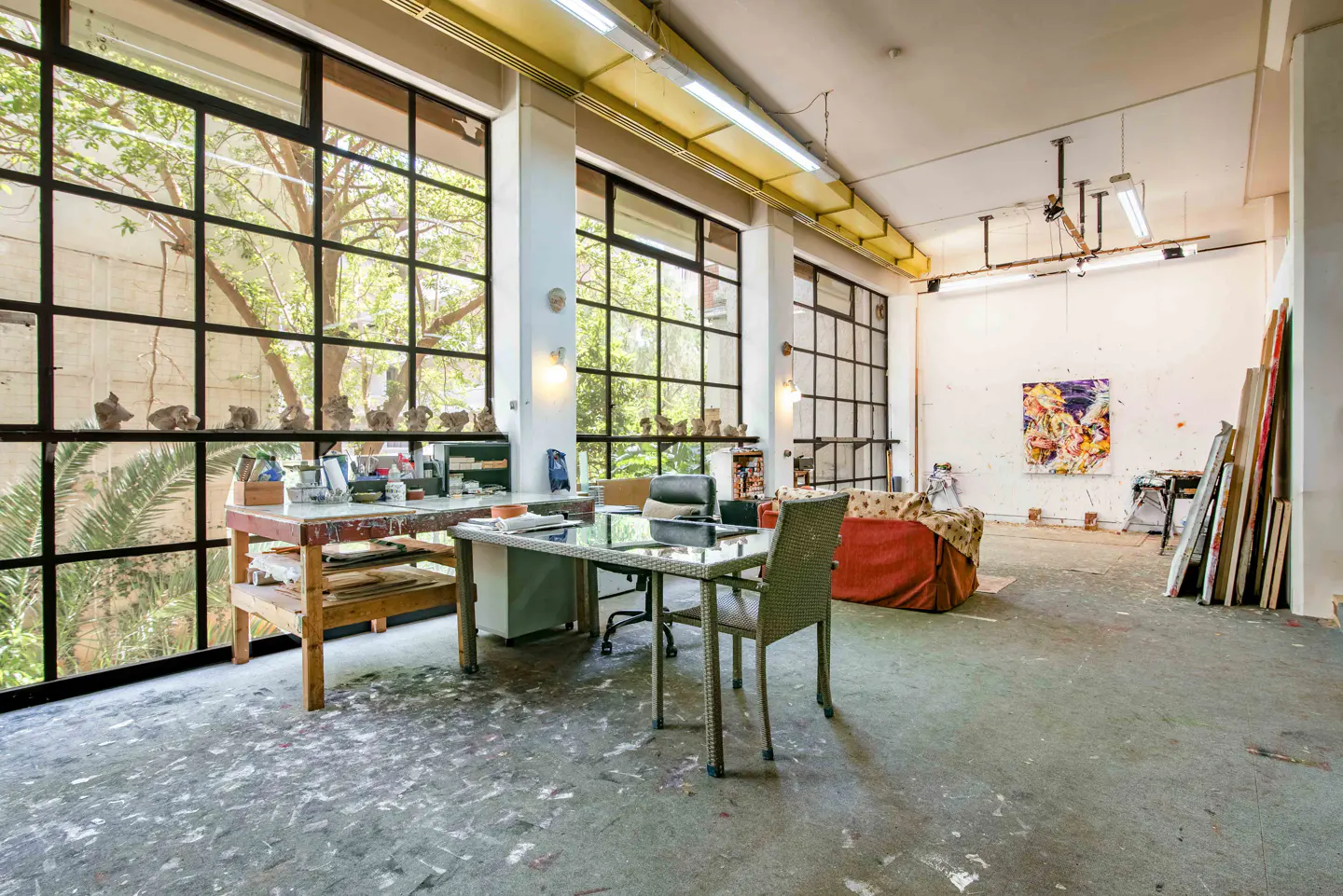 Bright artist's studio with large windows, paint-splattered floor, work tables, and a colorful abstract painting on the wall.