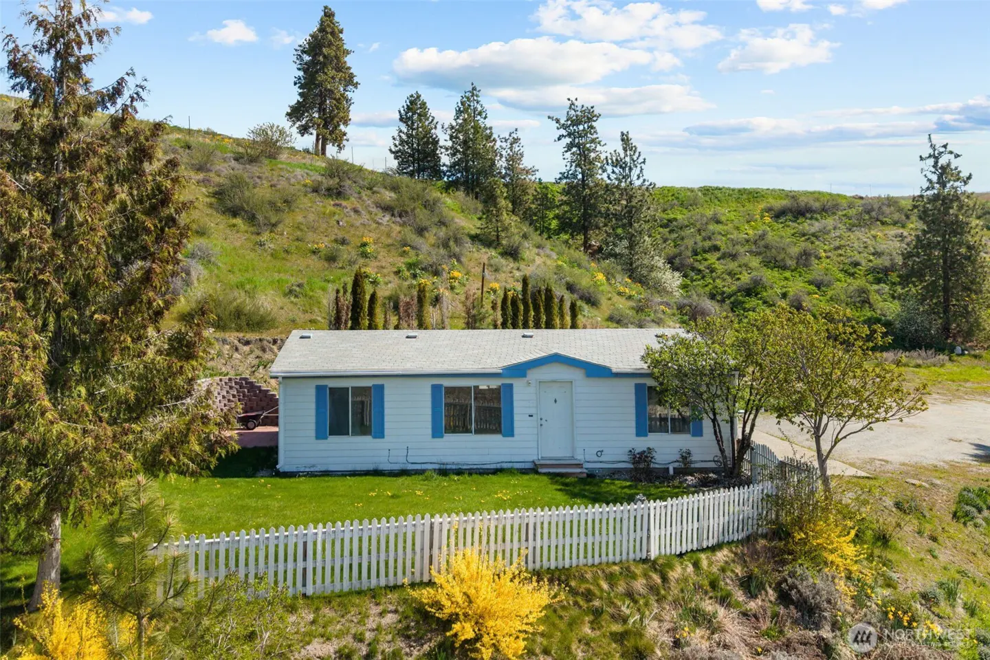 A white single-story house with blue shutters and a white picket fence on a grassy hill.