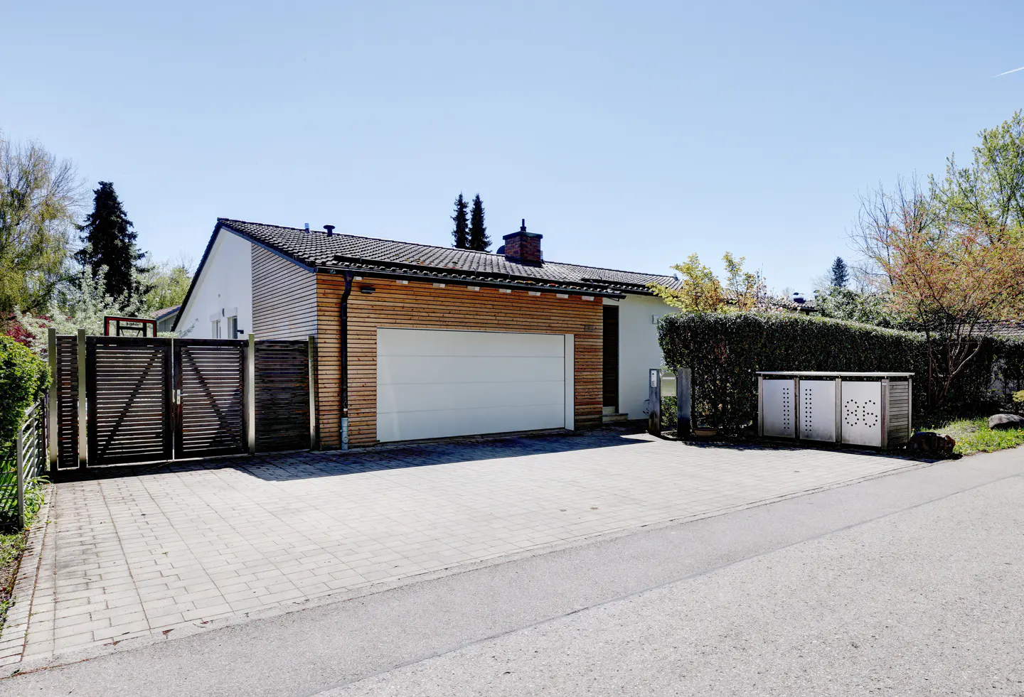 Exterior view of a modern single-story house with a white garage door, wood siding, and a gray brick driveway.