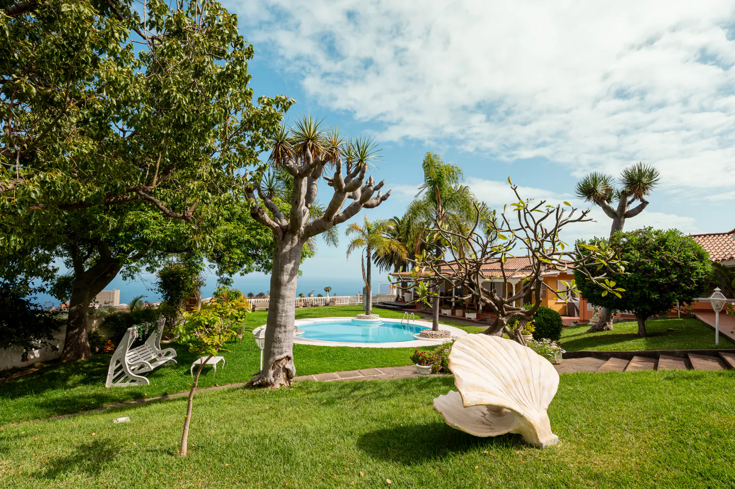 Lush green lawn with a pool, trees, and a large seashell sculpture. A white bench sits under a tree, with a house visible in the background.