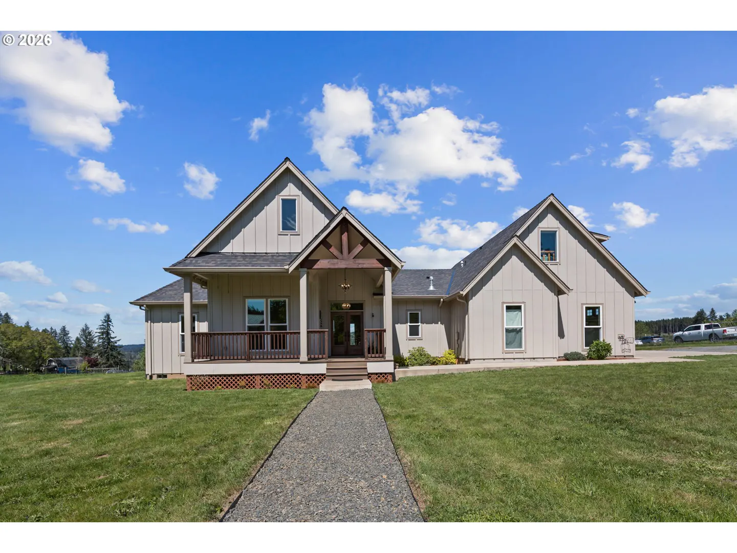 Beige farmhouse with a dark gray roof, a brown porch, and a gravel path on a green lawn under a blue sky.