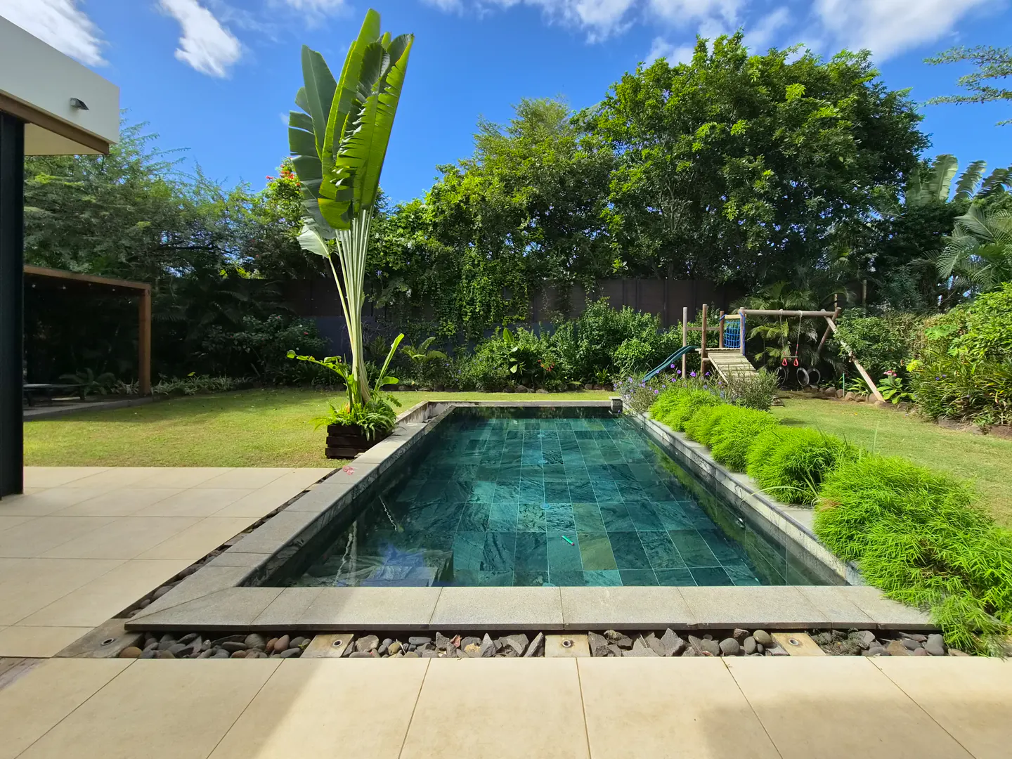 A backyard with a rectangular pool, green grass, trees, and a blue sky.