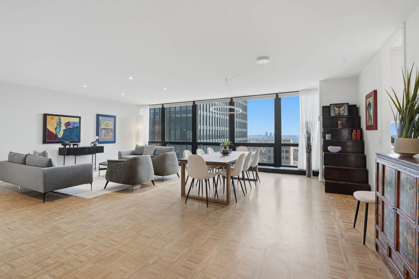 Bright, open-concept living and dining area with wood floors and city views. Gray sofas and chairs, white dining set, and modern art.