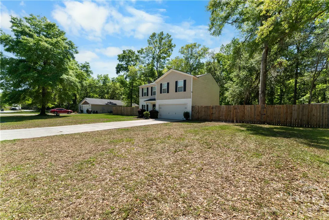 Two-story beige house with black shutters, a white garage door, and a long driveway. A wooden fence surrounds the backyard.