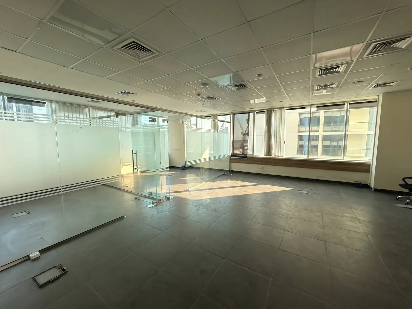 Empty office space with gray tile floor, glass-walled rooms, and large windows with sunlight streaming in.