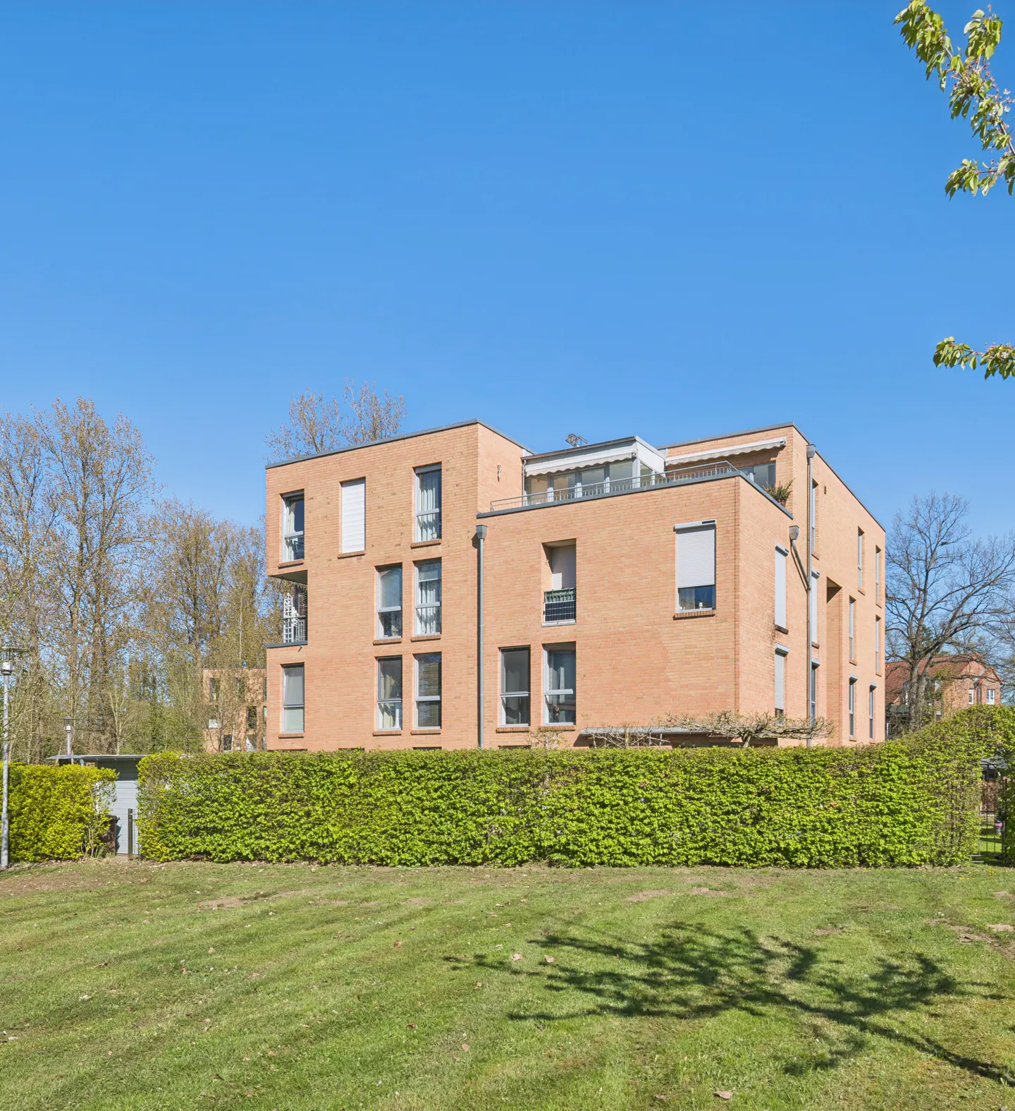 Modern brick apartment building with green lawn and trimmed hedges under a clear blue sky.