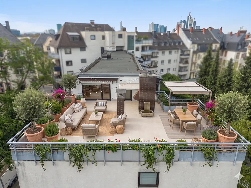 Aerial view of a rooftop terrace with beige furniture, potted plants, and a dining area. City buildings are visible in the background.