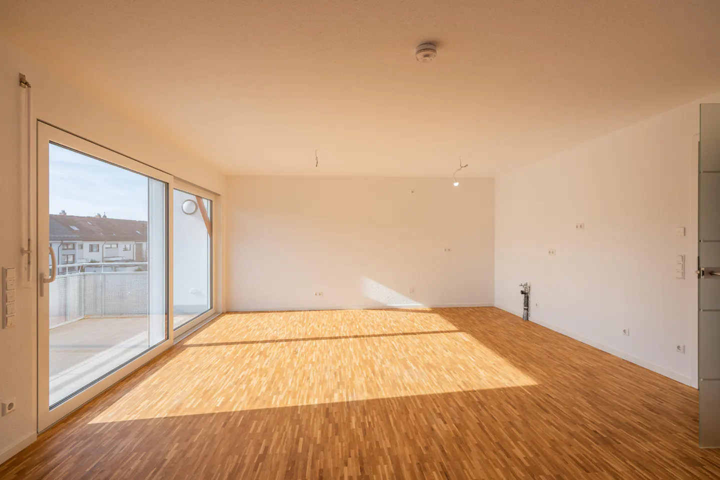 Bright, empty room with wood floors and white walls. Large sliding glass doors lead to a balcony with a view of houses.
