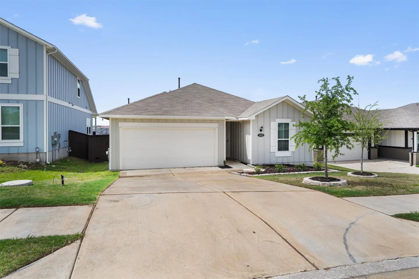 A one-story, light gray house with a white garage door and a concrete driveway under a blue sky.