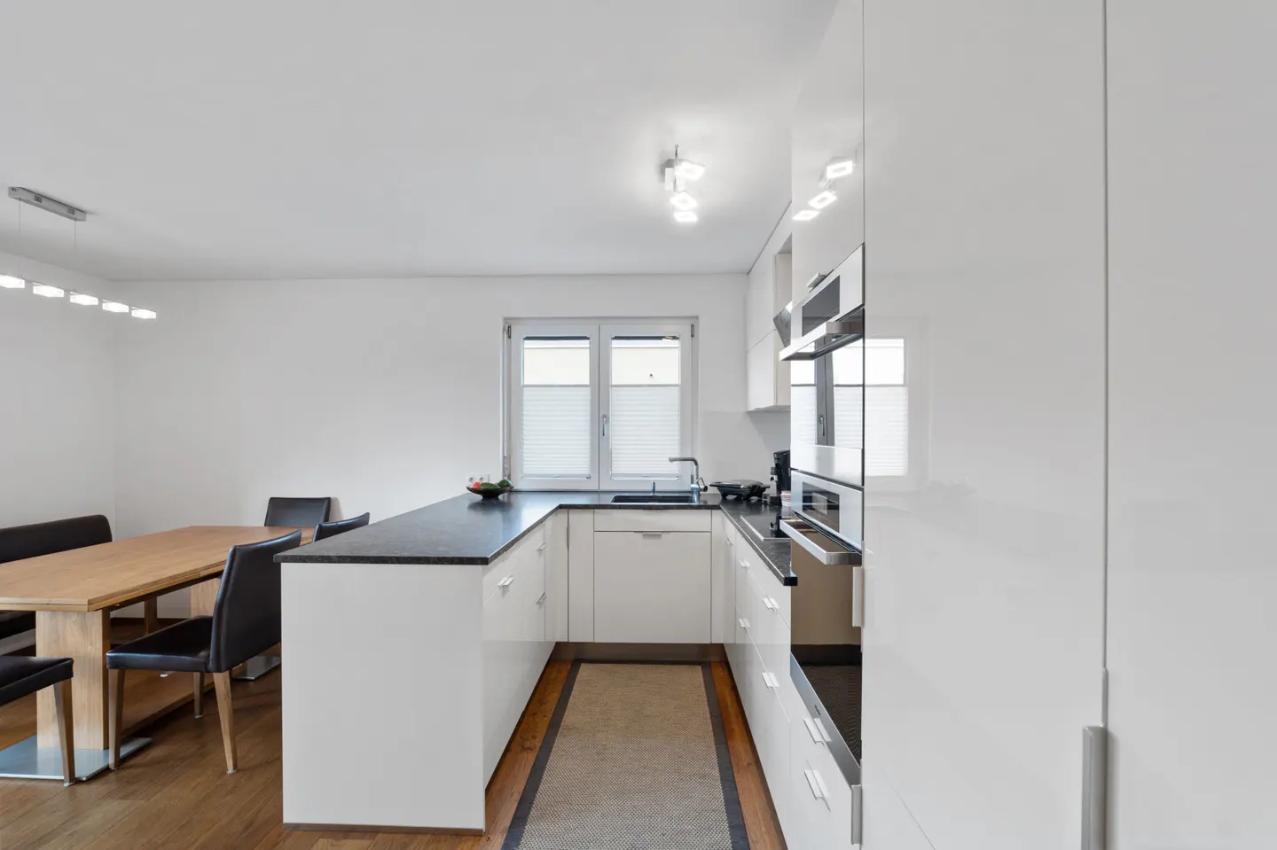 Bright, modern kitchen with white cabinets, black countertops, and stainless steel appliances. A wooden table and black chairs are visible.