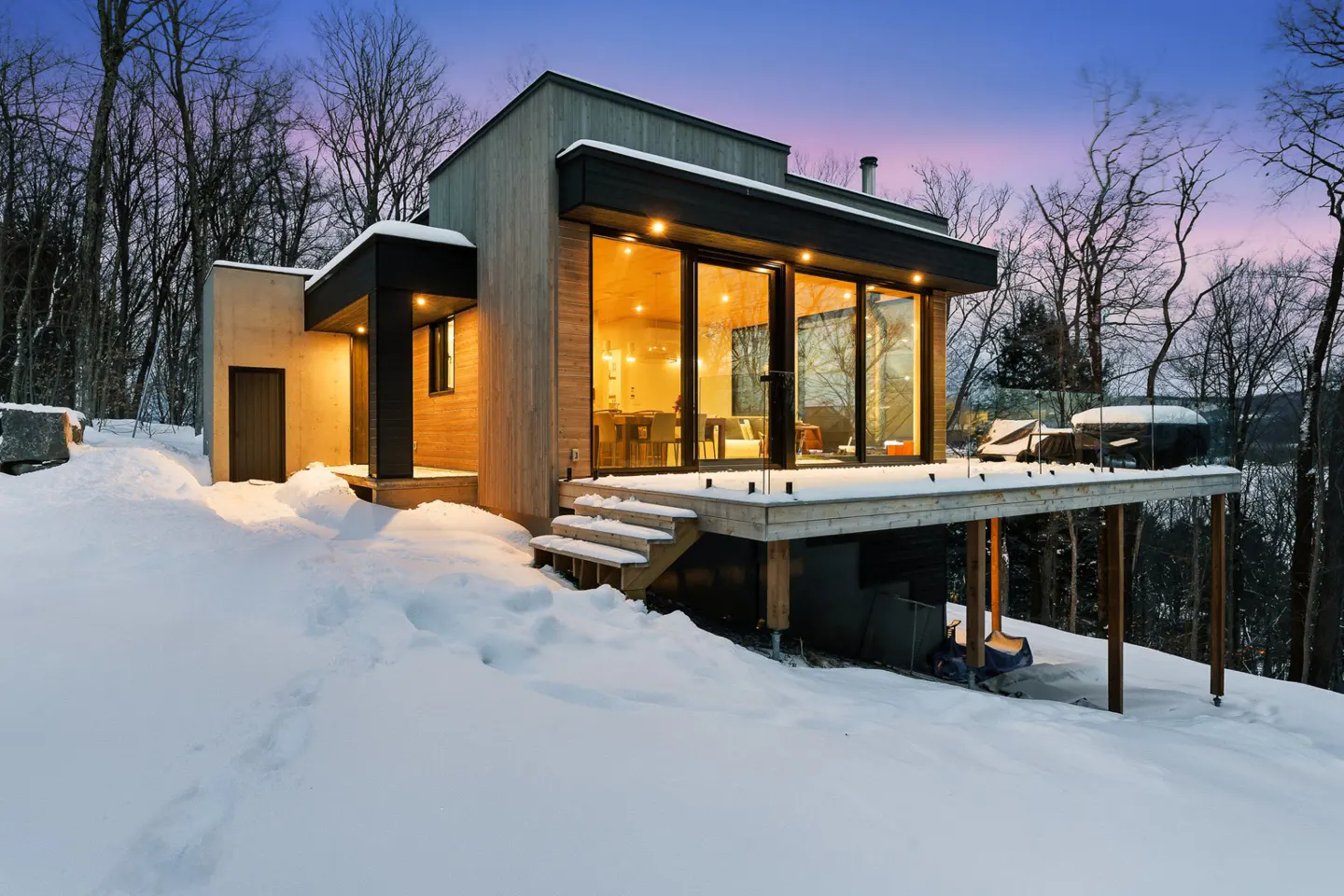 Modern home exterior in winter. The house has large windows, a deck, and is surrounded by snow and trees. The sky is a mix of blue and pink.