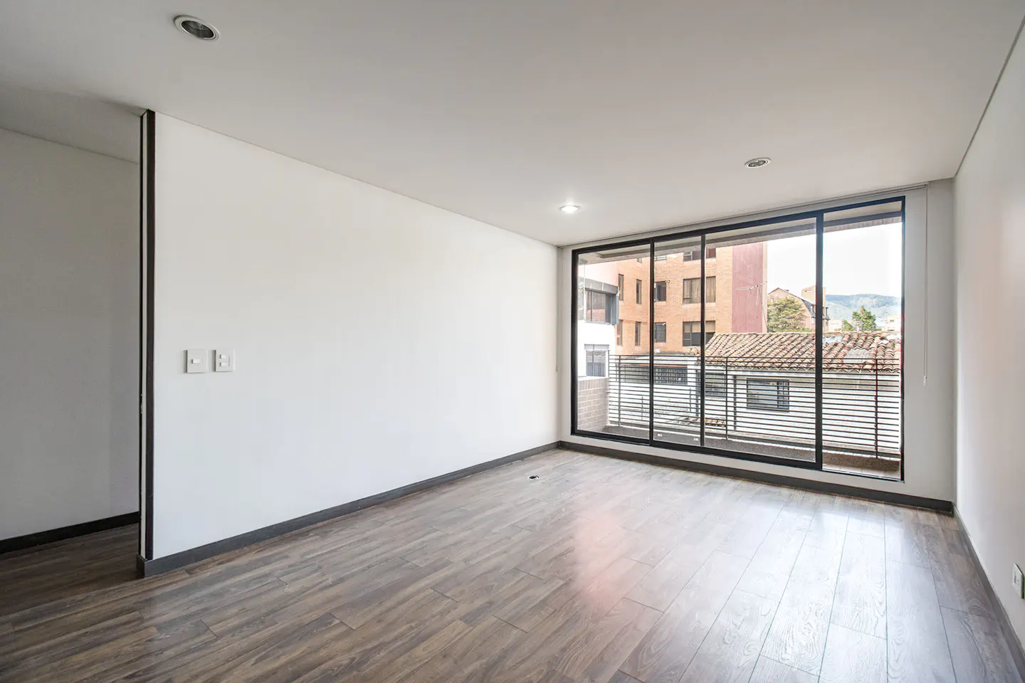 Bright, empty room with white walls and dark wood floors. Large black-framed windows offer a city view.