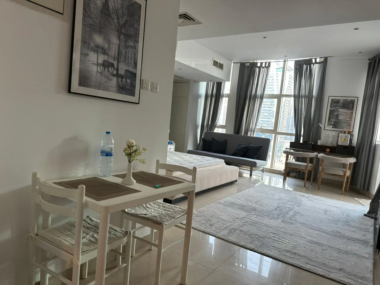 Modern studio apartment with a white dining set, a bed, a grey sofa, a desk with two chairs, and large floor-to-ceiling windows.