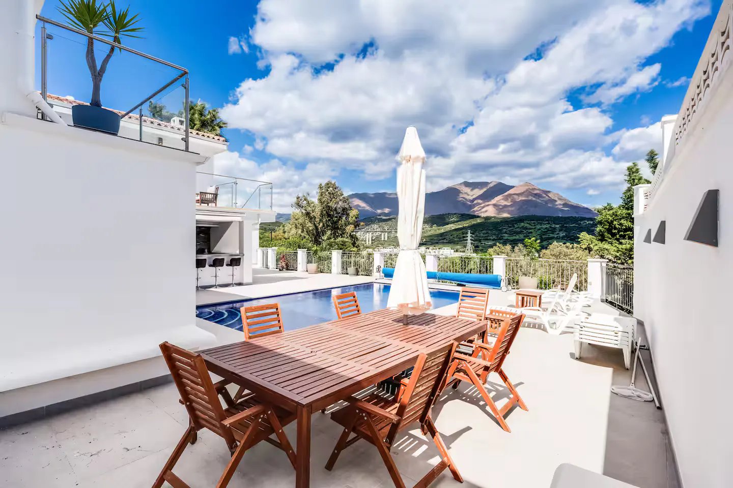 Outdoor patio with a wooden table and chairs, a pool, and mountain views under a cloudy sky.