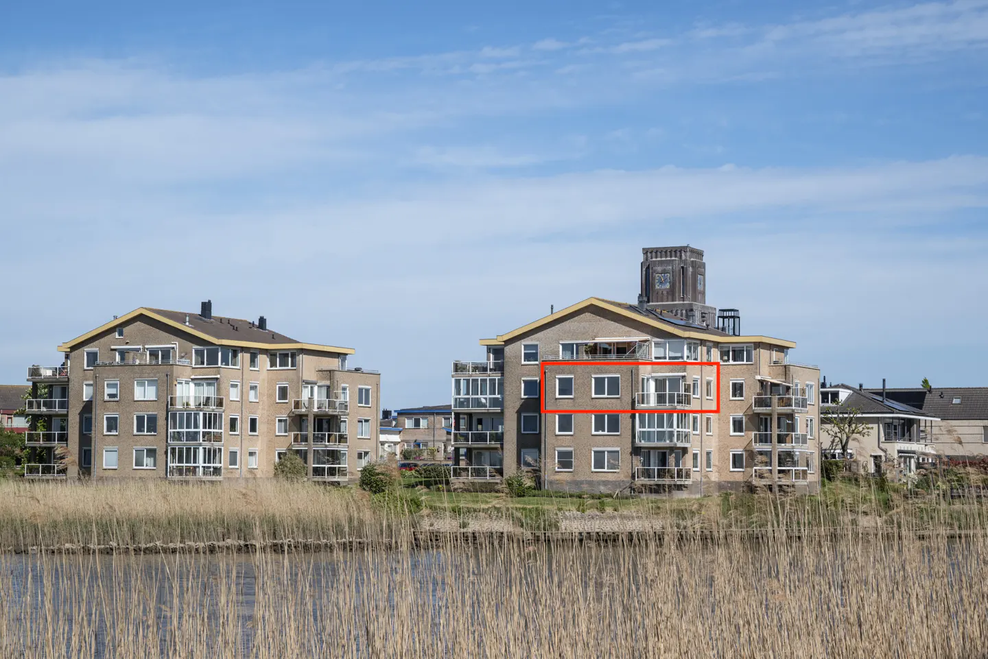Apartment building with balconies near water. The building is tan brick with white window frames. Tall grass in the foreground. Blue sky.