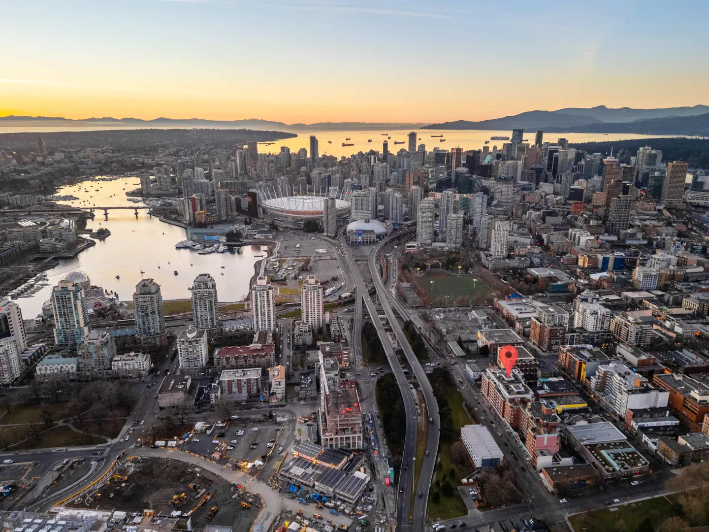 Aerial view of Vancouver, BC, at sunset. Stadium, skyscrapers, and waterways are visible. A red pin marks a building.