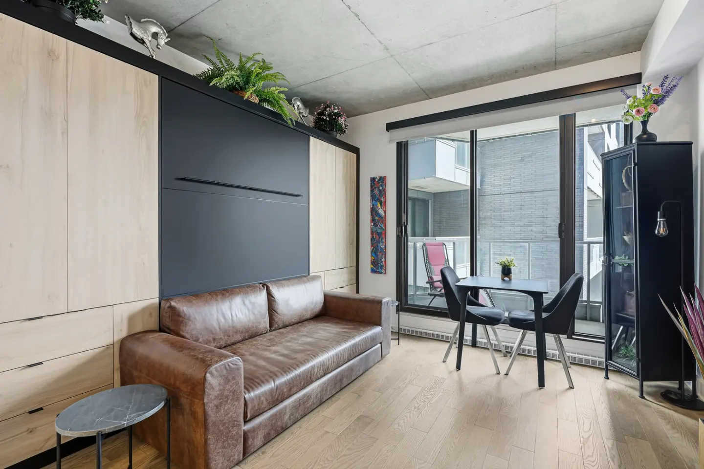 A modern apartment interior with a brown leather sofa in front of a black Murphy bed. A table and chairs sit near a balcony with a city view.