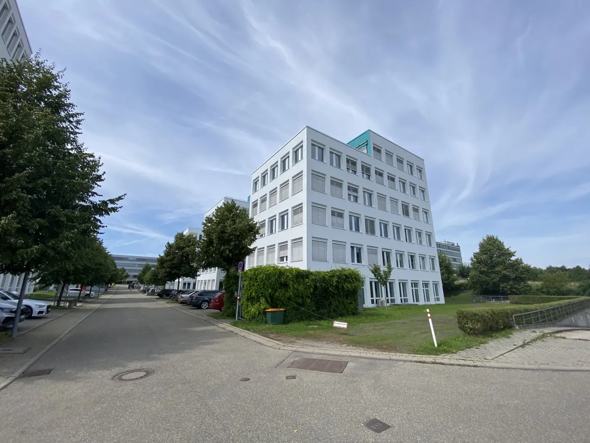Exterior view of a modern, white office building with many windows, green trees, and a gray paved road under a blue sky.